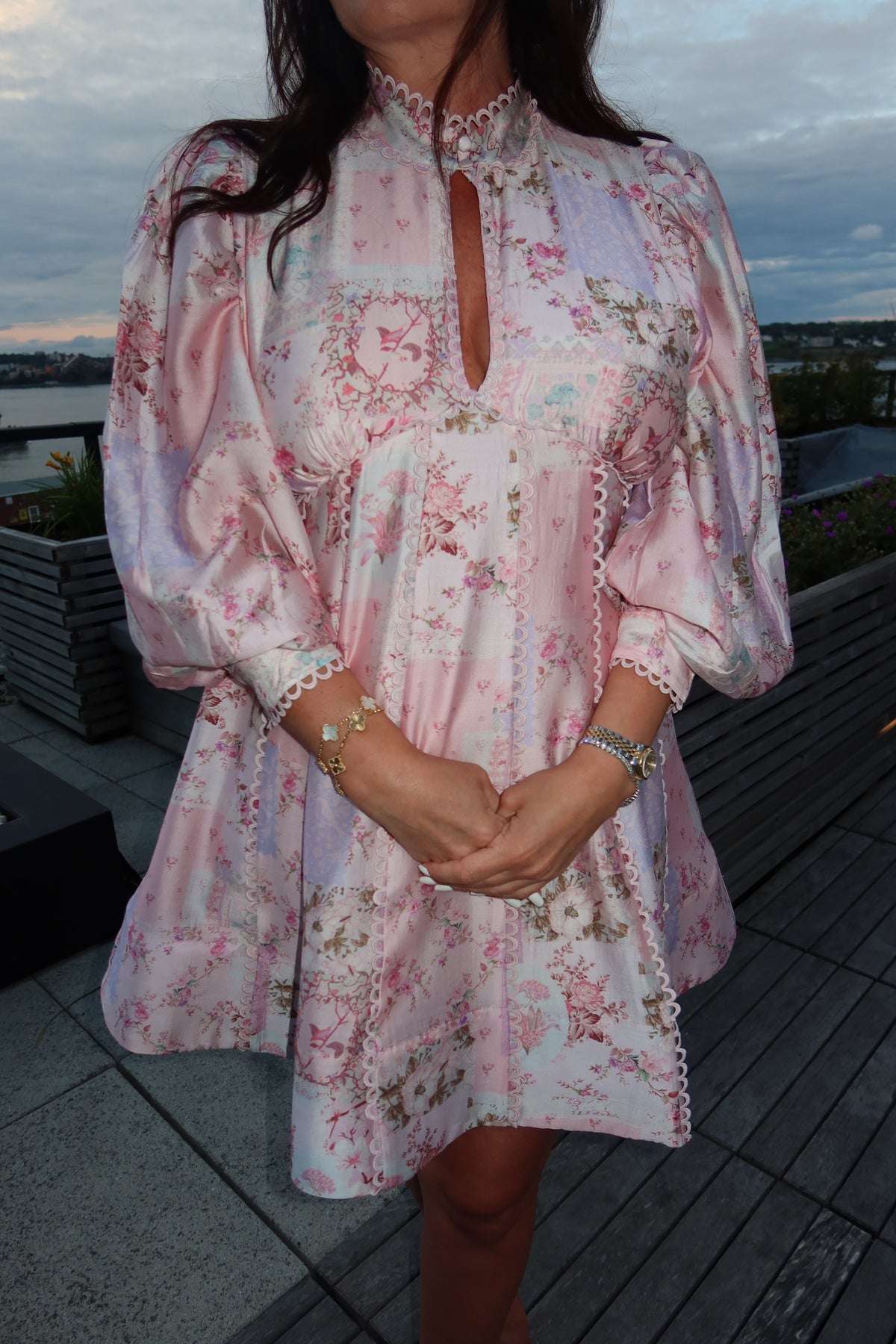 A woman wearing a light pink floral long-sleeve mini dress with a high mock neck and keyhole cutout, standing on a wooden deck at dusk.