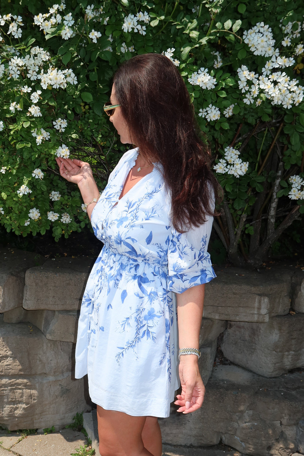 A woman wearing a light blue and white floral print mini dress stands outdoors in front of a stone wall and a green bush with white flowers.