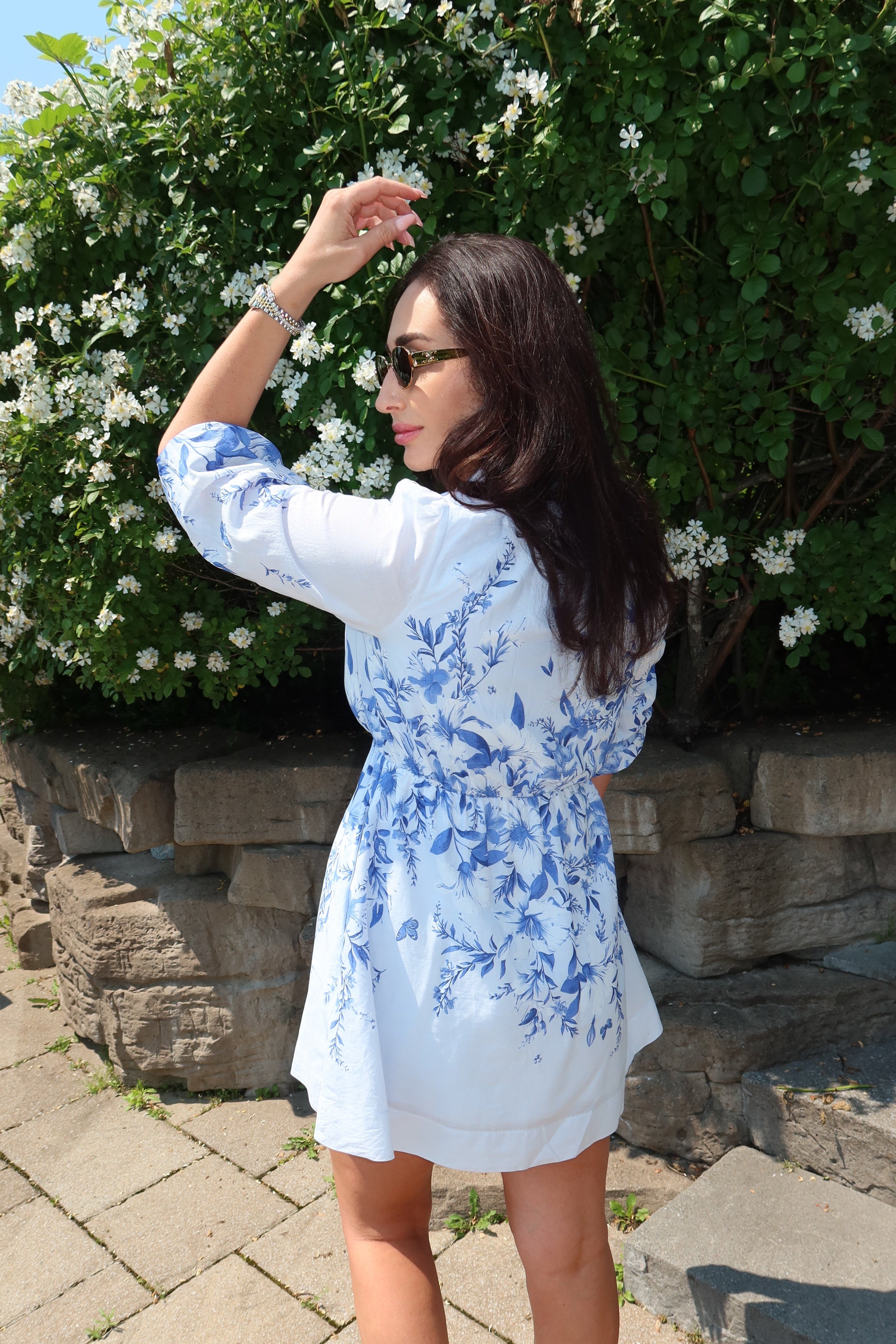 A woman wearing a white short-sleeve dress with a blue floral print stands outdoors in front of a stone wall and green flowering bushes. She is shown from the back and side, wearing sunglasses and white sandals.