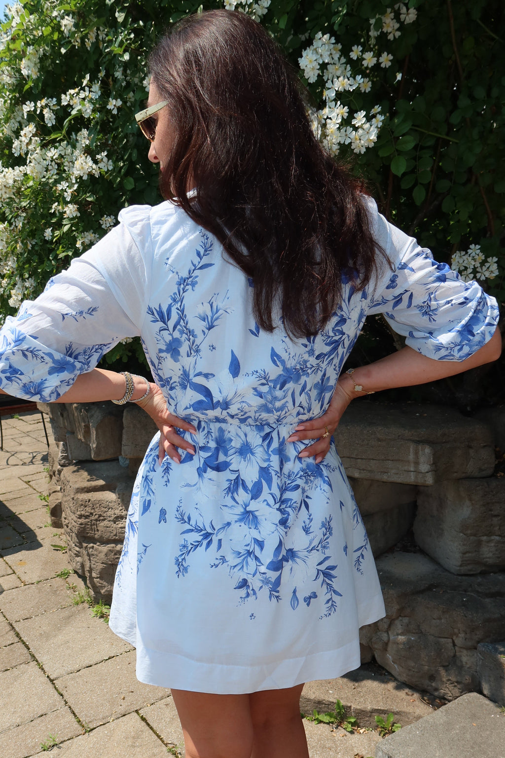 A woman wearing a white short-sleeve dress with a blue floral print, photographed from behind in an outdoor garden setting.