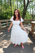 A woman wearing a white tiered midi dress with puff sleeves and pink sandals, standing on a paved path in a wooded outdoor setting.