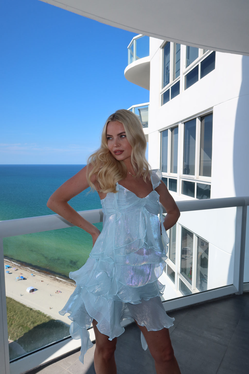 A blonde woman wearing a light blue ruffled dress stands on a balcony overlooking the ocean and a modern white building.