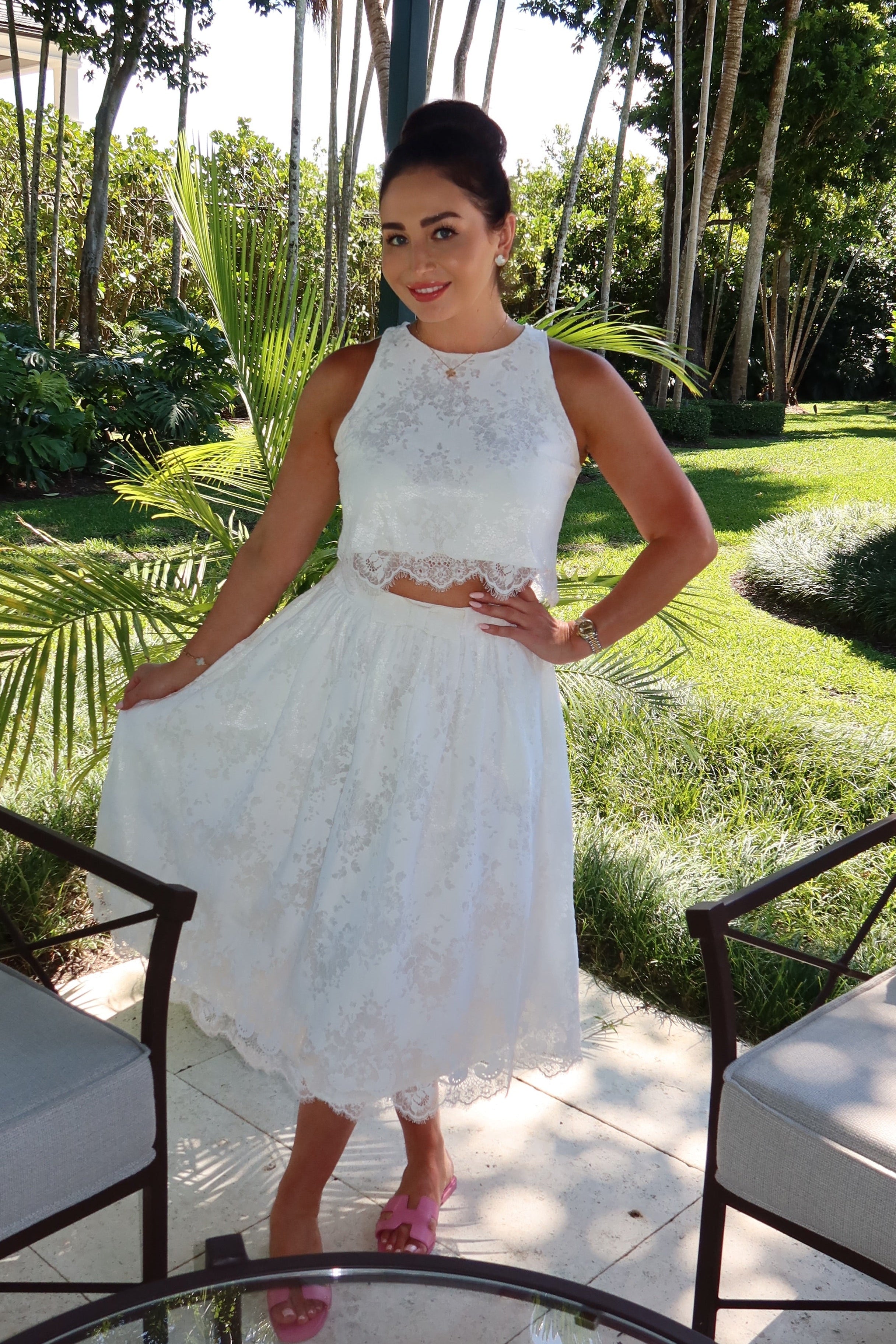 Woman in a white top and skirt standing in a garden setting with chairs and a table.