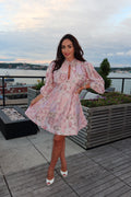 A woman posing on a rooftop terrace wearing a light pink floral print mini dress with long puff sleeves and white kitten heel sandals, with a city and water view in the background.