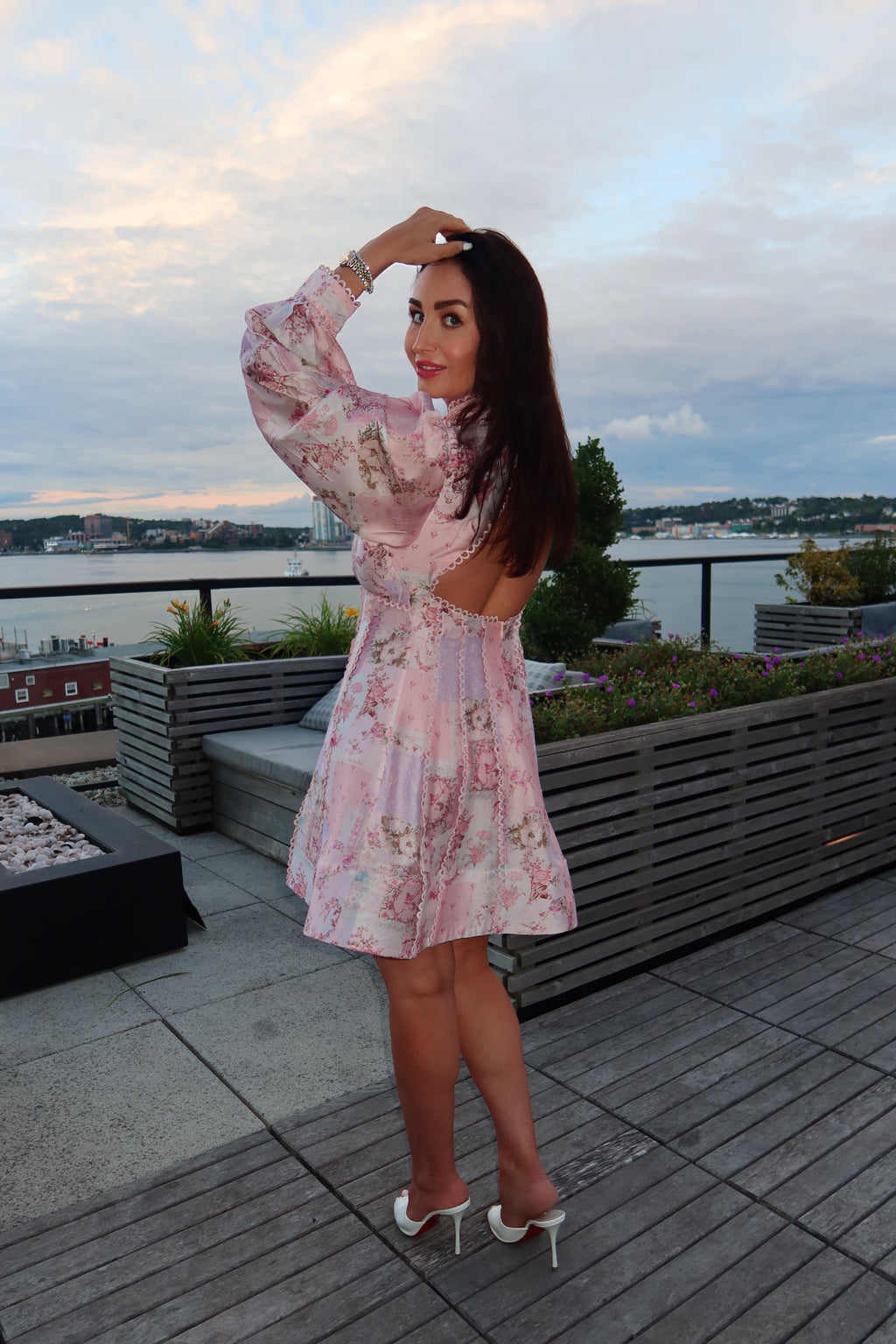 A woman stands on a rooftop terrace wearing a light pink floral mini dress with long puff sleeves and white heeled sandals. The background shows a city skyline and water under a cloudy sky.