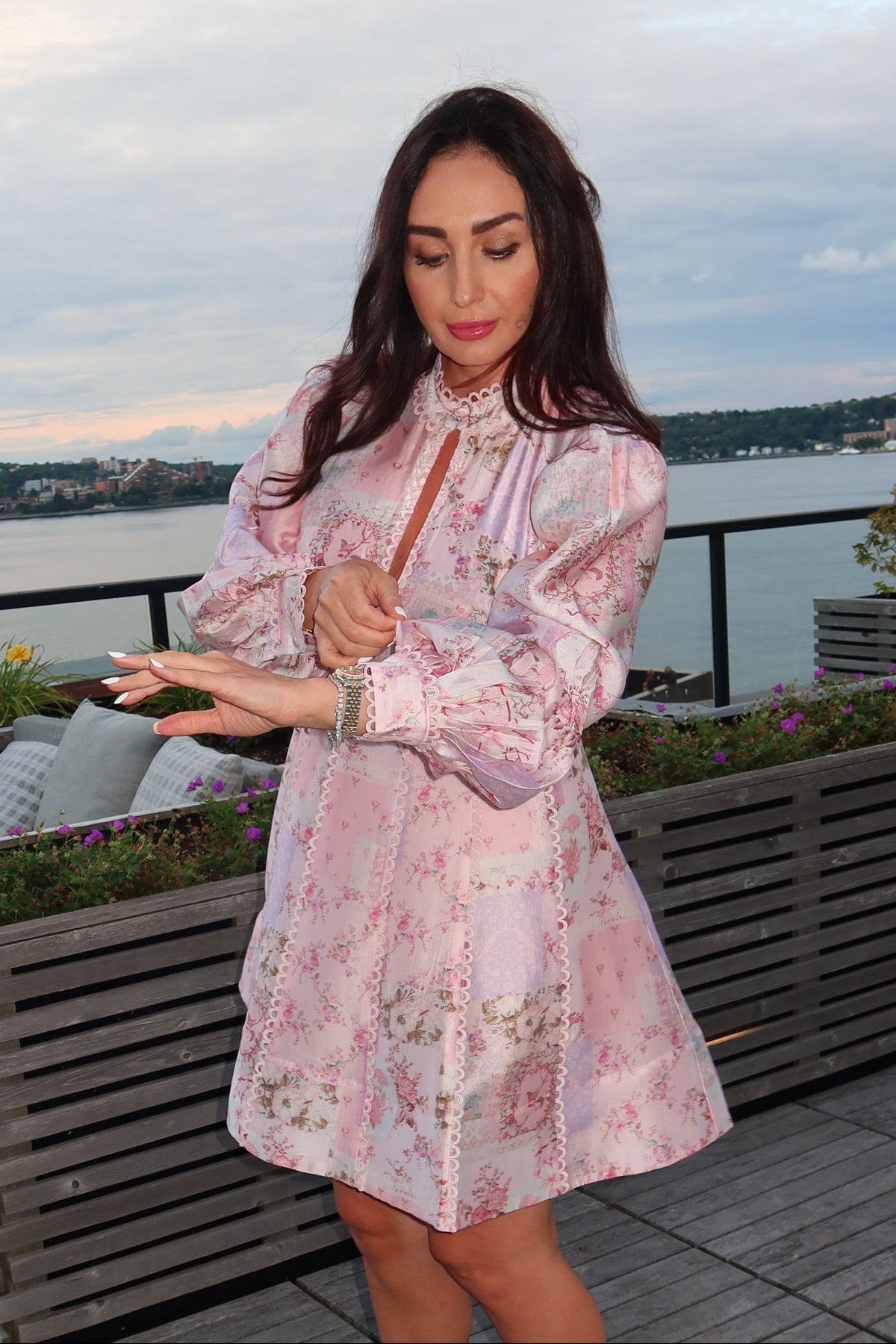 A woman stands on a rooftop terrace wearing a light pink floral long-sleeve dress with white high heels, with a lake and cloudy sky in the background.